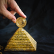 a finger holds a gold coin bitcoin on top of the pyramid on a black background.